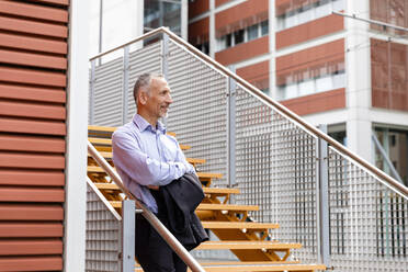 Thoughtful businessman with arms crossed leaning on railing at steps in city - EIF01342