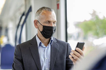 Männlicher Geschäftsmann, der ein Smartphone benutzt und einen Gesichtsschutz in der Straßenbahn während COVID-19 trägt - EIF01382
