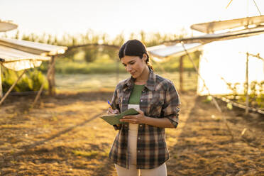 Junge Bäuerin schreibt in einem Notizblock, während sie auf einem Bauernhof steht - OIPF01078