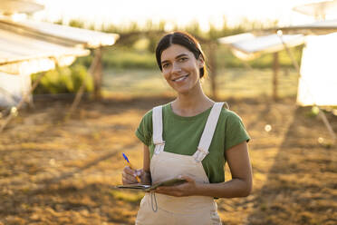 Lächelnde junge Bäuerin mit Notizblock auf einem Bauernhof an einem sonnigen Tag - OIPF01079