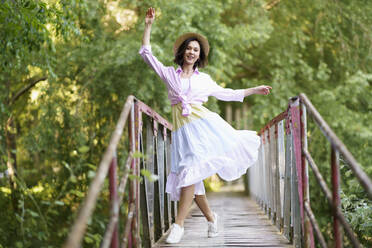 Young woman wearing hat dancing on footbridge in the countryside - JSMF02334