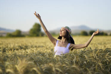 Schöne junge Frau mit Kopftuch steht mit erhobenen Armen in einem Weizenfeld - JSMF02346