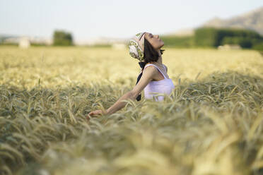 Junge Frau mit Kopftuch steht mit geschlossenen Augen in einem Weizenfeld - JSMF02348