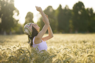 Junge Frau mit Kopftuch steht mit erhobenen Armen in einem Weizenfeld an einem sonnigen Tag - JSMF02349