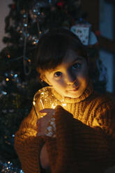 Nettes Mädchen im Pullover hält Weihnachtsbeleuchtung zu Hause - GEMF04842