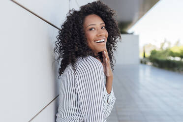 Cheerful woman with curly hair looking away while standing by wall - JRVF01042