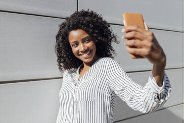 Smiling woman taking selfie through smart phone while standing by wall during sunny day - JRVF01054