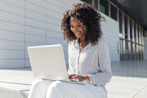 Lächelnde junge Geschäftsfrau, die einen Laptop benutzt, während sie vor einem Bürogebäude sitzt - JRVF01056