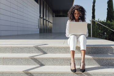 Berufstätige Frau, die einen Laptop benutzt, während sie an einem sonnigen Tag auf einer Treppe sitzt - JRVF01057