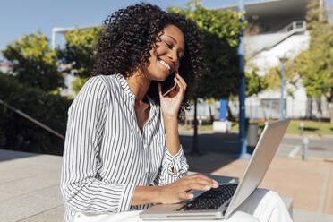 Lächelnde weibliche Fachkraft, die sich mit einem Smartphone unterhält, während sie an einem sonnigen Tag mit einem Laptop im Büropark sitzt - JRVF01061