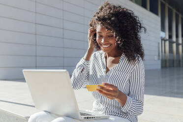 Lächelnde Frau mit Kreditkarte beim Online-Shopping, während sie vor einem Bürogebäude sitzt - JRVF01063