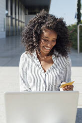 Glückliche junge Geschäftsfrau mit Laptop beim Online-Shopping vor einem Bürogebäude - JRVF01065
