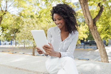 Smiling curly haired businesswoman using digital tablet while sitting at office park - JRVF01069