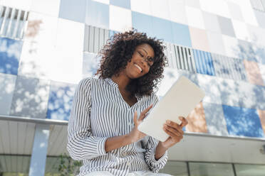 Cheerful curly haired businesswoman using digital tablet while sitting in front of office building - JRVF01073