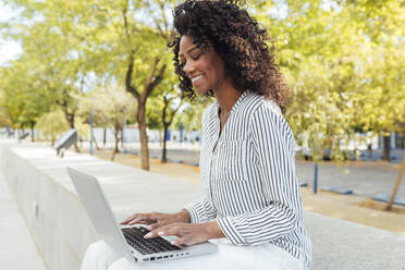 Smiling curly haired businesswoman typing while using laptop sitting at office park - JRVF01082