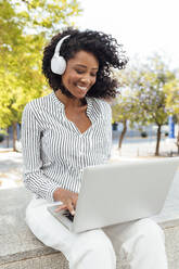 Smiling female professional listening music through headphones while using laptop sitting at office park - JRVF01083