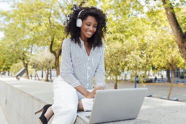 Smiling female professional listening music through headphones while sitting with laptop at office park - JRVF01085