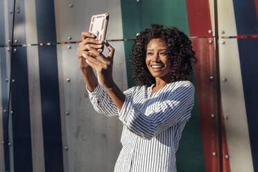 Glückliche junge Frau, die ein Selfie mit ihrem Smartphone macht, während sie vor einer Wand steht - JRVF01096