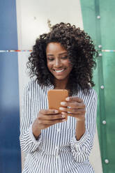 Smiling curly haired woman text messaging through smart phone while standing in front of wall - JRVF01098