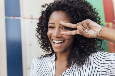 Cheerful woman winking eye while gesturing peace sign standing in front of wall - JRVF01100