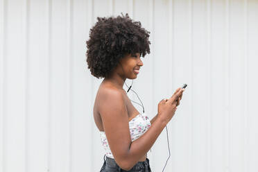 Smiling Afro woman with smart phone listening music through headphones by white wall - JRVF01115