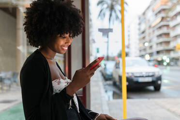 Lächelnde junge Frau, die an der Bushaltestelle ein Mobiltelefon benutzt - JRVF01128