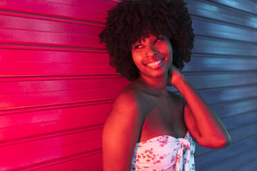 Smiling Afro woman with hand in hair looking away while standing in front of shutter at night - JRVF01133