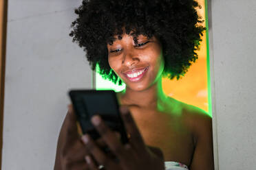 Young Afro woman smiling while using smart phone in front of neon lights - JRVF01136
