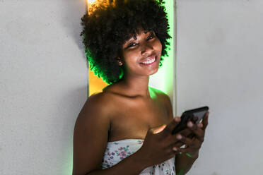 Lächelnde Afro-Frau mit Mobiltelefon vor Neonlicht - JRVF01137