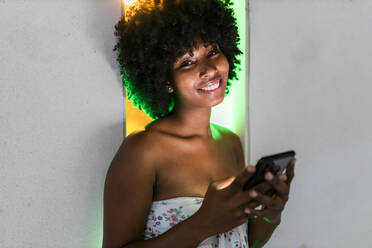 Smiling Afro woman with mobile phone standing in front of neon lights - JRVF01137