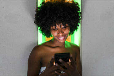 Young Afro woman smiling while texting message in front of neon lights - JRVF01138