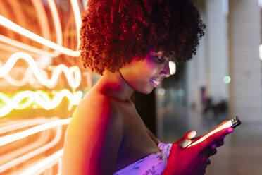 Afro-Frau, die eine Textnachricht auf einem Mobiltelefon bei Neonlichtern schreibt - JRVF01143