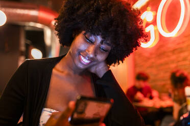 Lächelnde Afro-Frau, die eine SMS über ihr Smartphone in einer Bar schreibt - JRVF01146