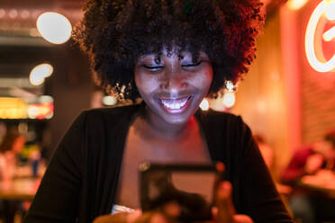 Afro-Frau lächelt bei der Benutzung eines Mobiltelefons in einer Bar - JRVF01147