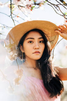 From below peaceful thoughtful ethnic female in straw hat and dress standing under blooming fragrant flowers on tree branches in orchard looking at camera - ADSF24729
