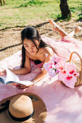 Ethnische asiatische Frau in stilvollem Kleid auf einer Decke liegend mit Blumen in einem Weidenkorb und Schuhen mit Strohhut und einem Buch lesend im Garten - ADSF24737