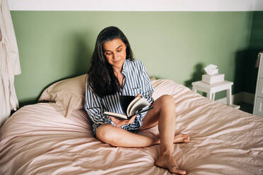 Entspannte reife hispanische Frau in Freizeitkleidung sitzt im Bett und liest ein interessantes Buch - ADSF24745