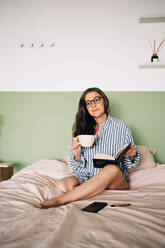 Kluge Frau mit Brille, langen Haaren und Notizbuch, die wegschaut und sich auf einem weichen Bett im Schlafzimmer ausruht - ADSF24747
