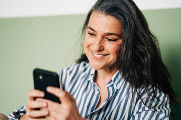 Positive hispanische Frau mittleren Alters mit langen dunklen Haaren in Freizeitkleidung, die lächelt, während sie auf dem Bett zu Hause sitzt und mit ihrem Handy Nachrichten austauscht - ADSF24748