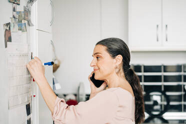 Side view of positive middle aged female making notes in calendar on fridge while having phone call in modern kitchen - ADSF24754