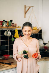 Lächelnde Frau mittleren Alters mit rotem Glockenpapier Lauch und Gurken Blick nach unten, während stehend in der Nähe von Küchentisch mit Geschirr - ADSF24756
