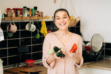 Lächelnde Frau mittleren Alters mit rotem Glockenpapier, Lauch und Gurken, die in die Kamera schaut, während sie in der Nähe der Küchentheke mit Geschirr steht - ADSF24757