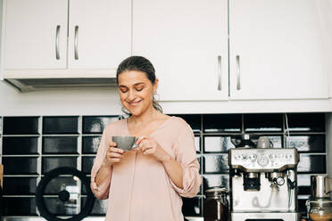 Fröhliche Frau mittleren Alters mit einer Tasse Heißgetränk an der Küchentheke mit weißen Schränken und moderner Kaffeemaschine zu Hause - ADSF24760