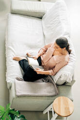 From above full body of barefoot female reading book while sitting on comfortable sofa in living room with green plant at home - ADSF24763