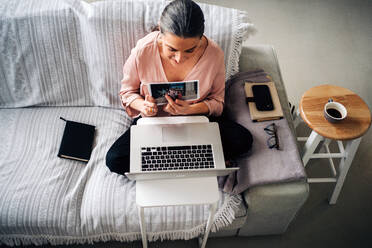 Von oben von einer Frau, die auf dem Sofa sitzt und ein Bild zeigt, während sie einen Videochat über einen Laptop im Wohnzimmer zu Hause führt - ADSF24771