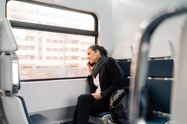 Side view of content female sitting on passenger seat and looking out window while having phone call in modern train - ADSF24775