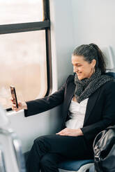 Side view of positive middle aged female with scarf taking picture on cellphone while sitting on passenger seat near window in wagon during journey - ADSF24777