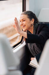 Seitenansicht einer positiven Frau, die mit der Hand winkt und aus dem Fenster schaut, während sie auf dem Beifahrersitz eines Wagens sitzt - ADSF24778