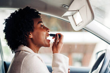 Side view of African American female driver sitting in automobile and applying lipstick while looking at mirror - ADSF24782