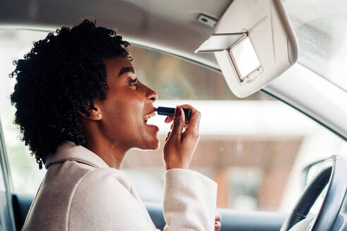 Side view of African American female driver sitting in automobile and applying lipstick while looking at mirror - ADSF24782